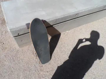 High-angle photo of a skateboard resting against a concrete ledge with a long shadow across the ground